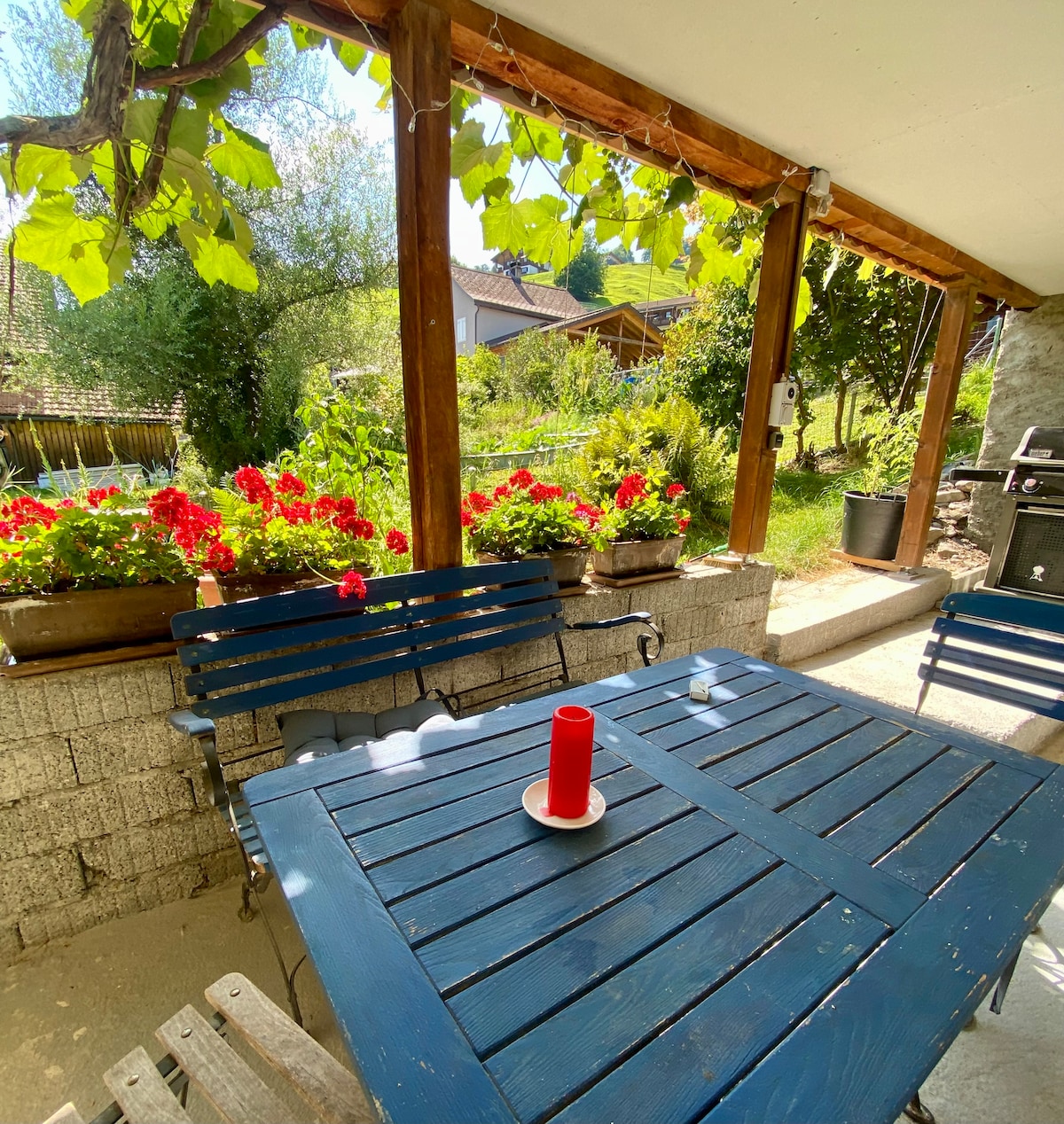 An outdoor seating area is partially shaded by a wooden roof. A blue table is surrounded by slatted benches and features a red candle in the center. Flower boxes filled with vibrant red blossoms enhance the space, accompanied by greenery in the background.
