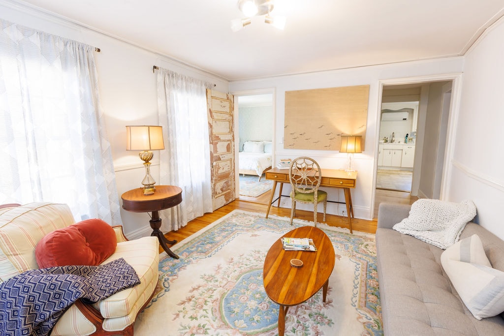 A cozy living room features a light color palette with large windows allowing natural light. The space includes a comfortable sofa, a round wooden coffee table, and a desk with an antique chair. Textiles in soft tones and subtle patterns add warmth.