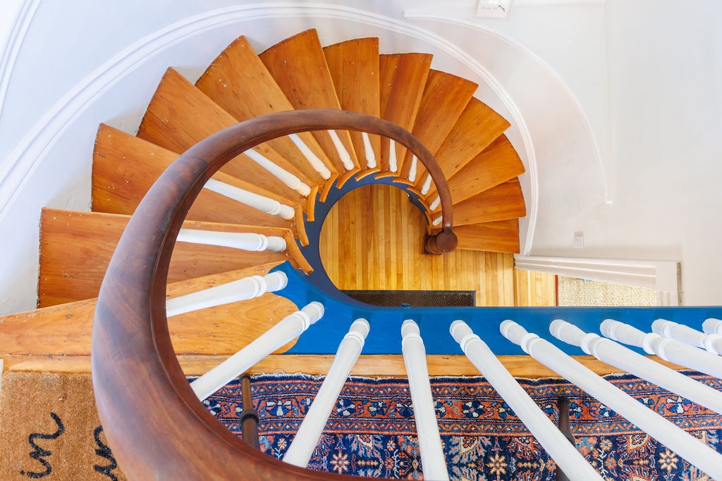 A spiral wooden staircase is viewed from above, showcasing smooth, polished steps that curve gracefully. White balusters line the staircase, and a colorful area rug is visible at the base, adding warmth to the space.
