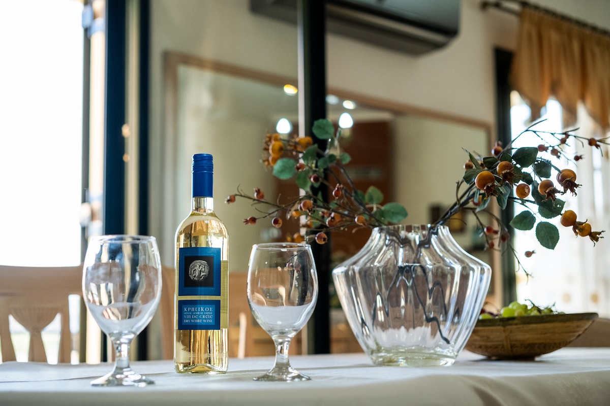 A dining table is set with two clear wine glasses positioned beside a bottle of white wine, prominently displayed. A transparent glass vase holds an arrangement of twigs with small leaves and berries, adding a natural touch to the setting.