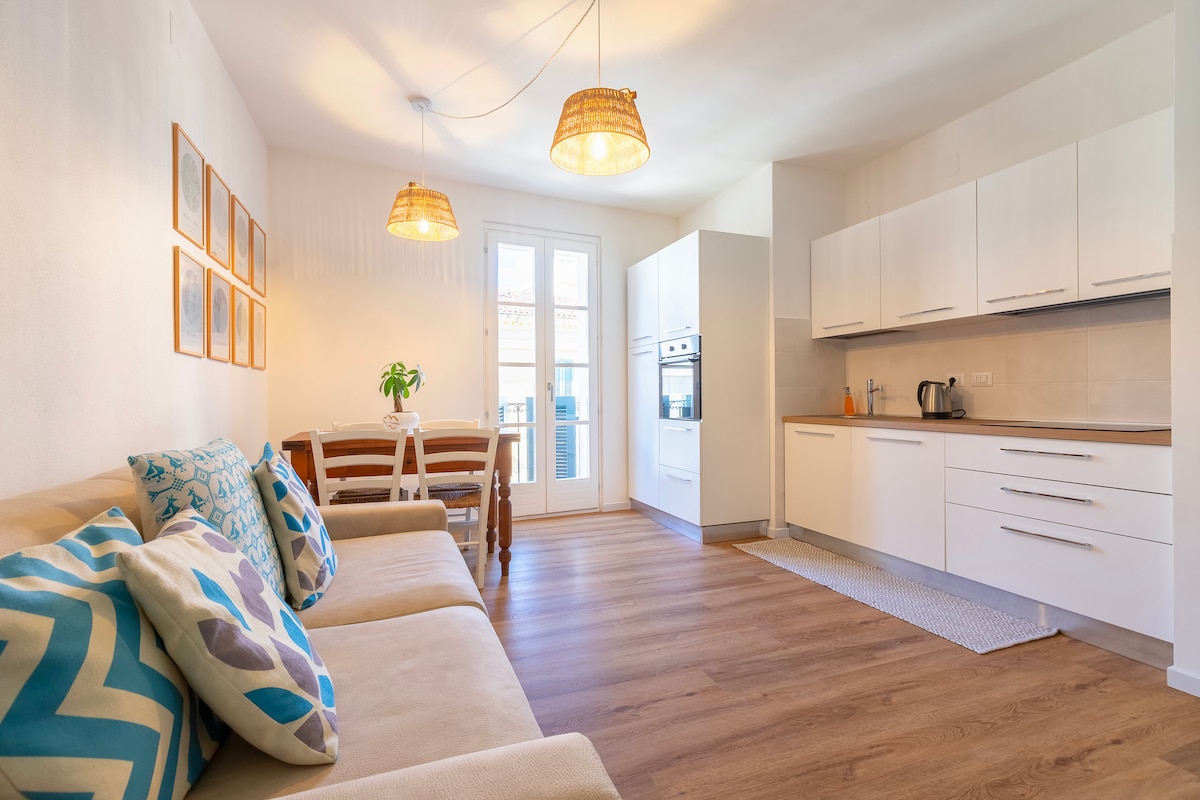 A bright living space features a modern kitchen area with white cabinetry, complemented by a light wood floor. A comfortable seating area includes a sofa adorned with decorative cushions. Two pendant lights hang from the ceiling, providing illumination over the dining table nearby.