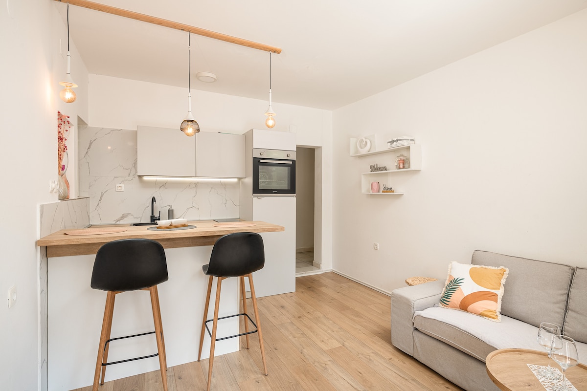 A modern kitchen area features sleek cabinetry and a marble backsplash, complemented by warm pendant lighting. Two wooden bar stools are situated at a counter, while a comfortable sofa is positioned nearby. The open layout invites a relaxed atmosphere.