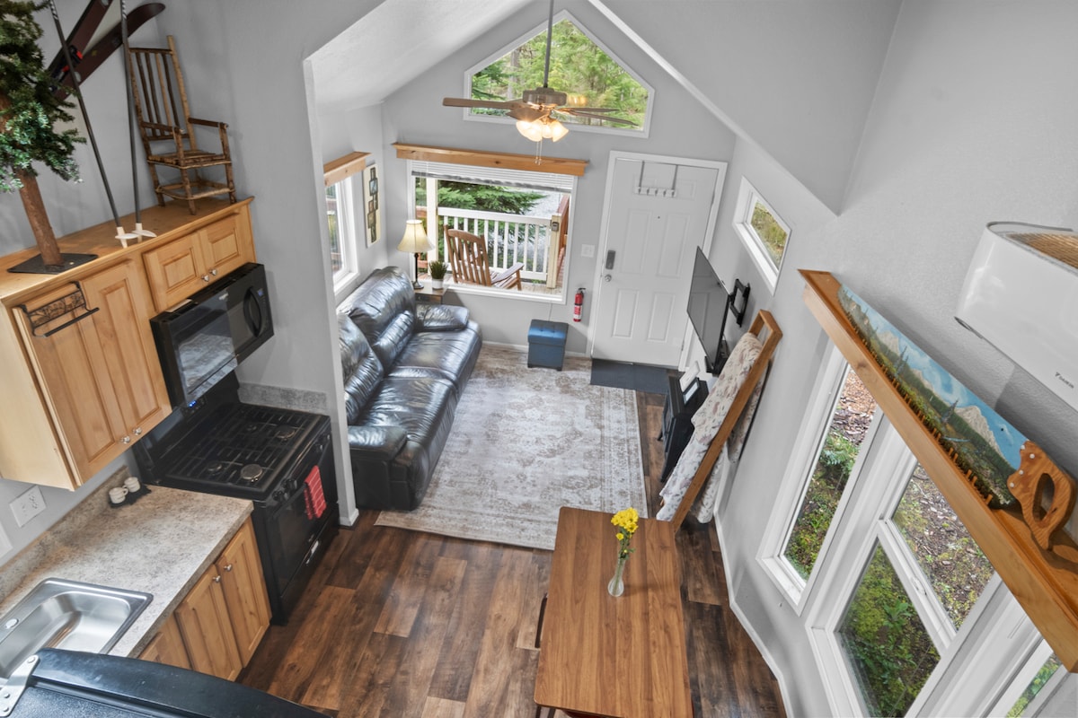 An interior view showcases an open living area with a cozy seating arrangement, a dining table, and a fully equipped kitchen. Large windows allow natural light to fill the space, providing a view of the surrounding greenery.