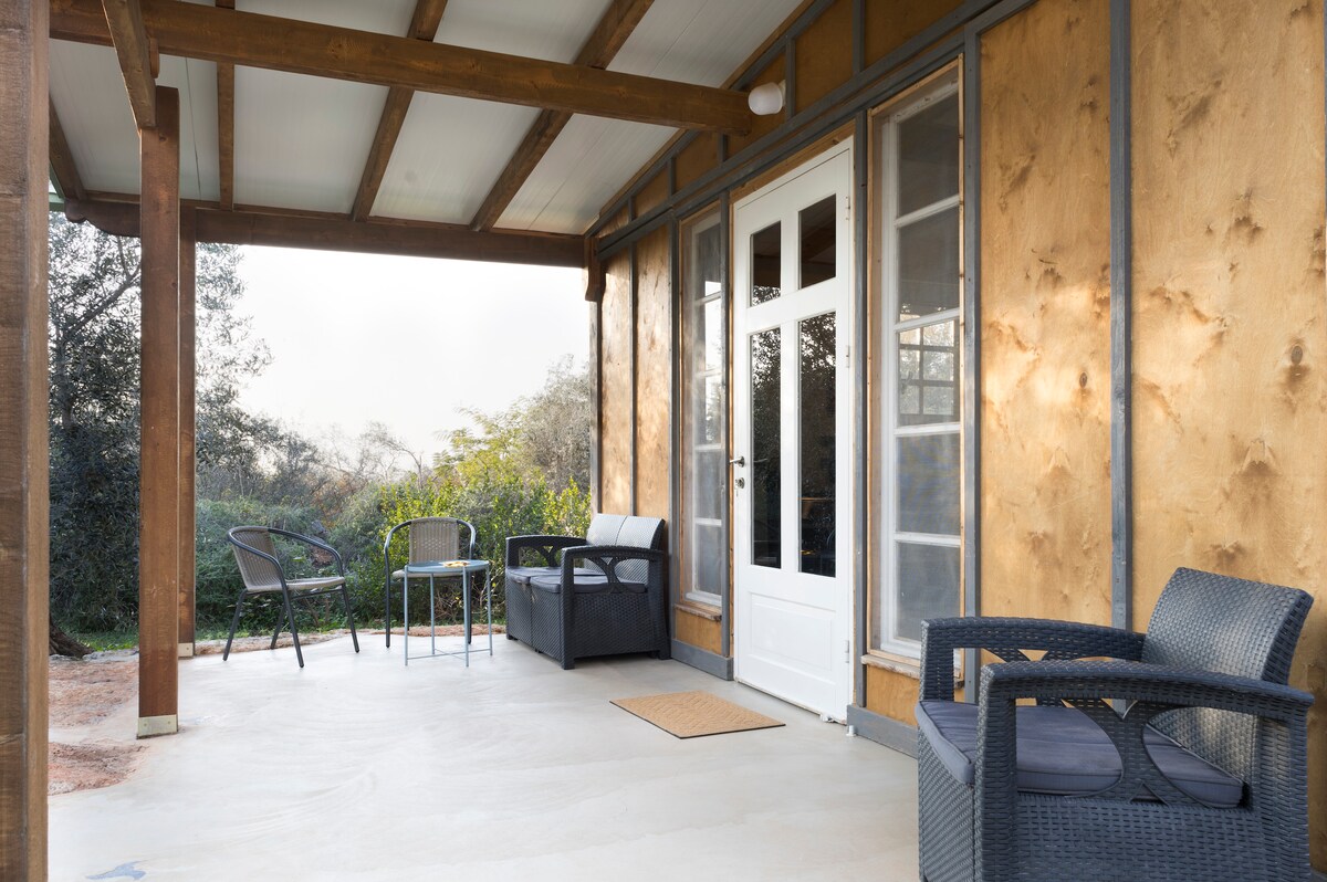 A spacious porch features comfortable seating options, including two wicker chairs and a small table. Natural light filters through large windows, inviting the outdoors inside, while the surrounding greenery enhances the peaceful atmosphere of this tranquil retreat.
