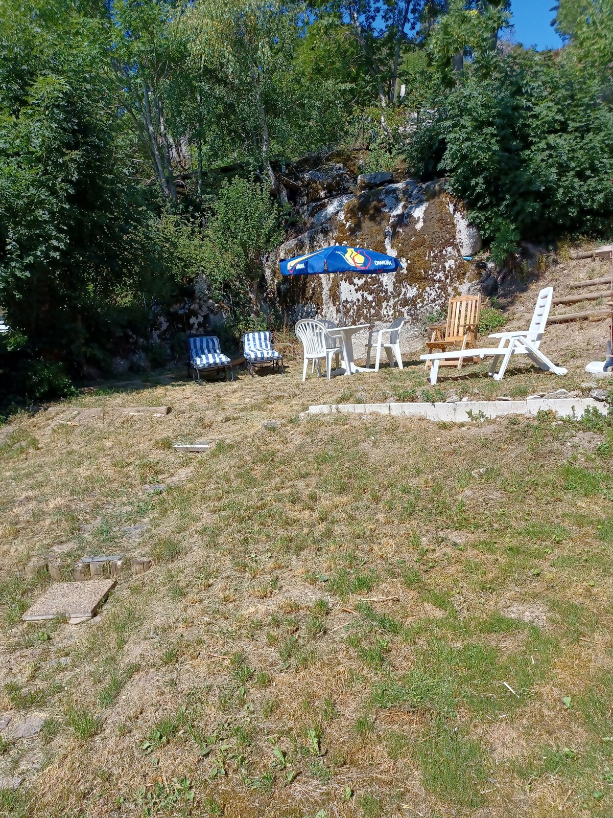 A serene outdoor area is captured, featuring a small seating arrangement with four chairs and a table. A colorful umbrella provides shade, while a rocky backdrop is framed by greenery. The space offers a peaceful setting for relaxation in nature.