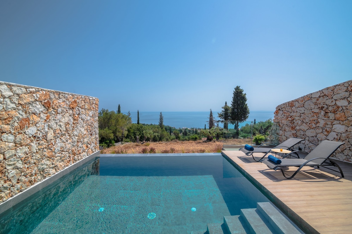 A serene infinity pool reflects the blue sky above, bordered by natural stone walls. Lounge chairs are positioned along the poolside, offering a tranquil space to relax. The expansive view showcases the sea and distant greenery, creating a peaceful and captivating setting.