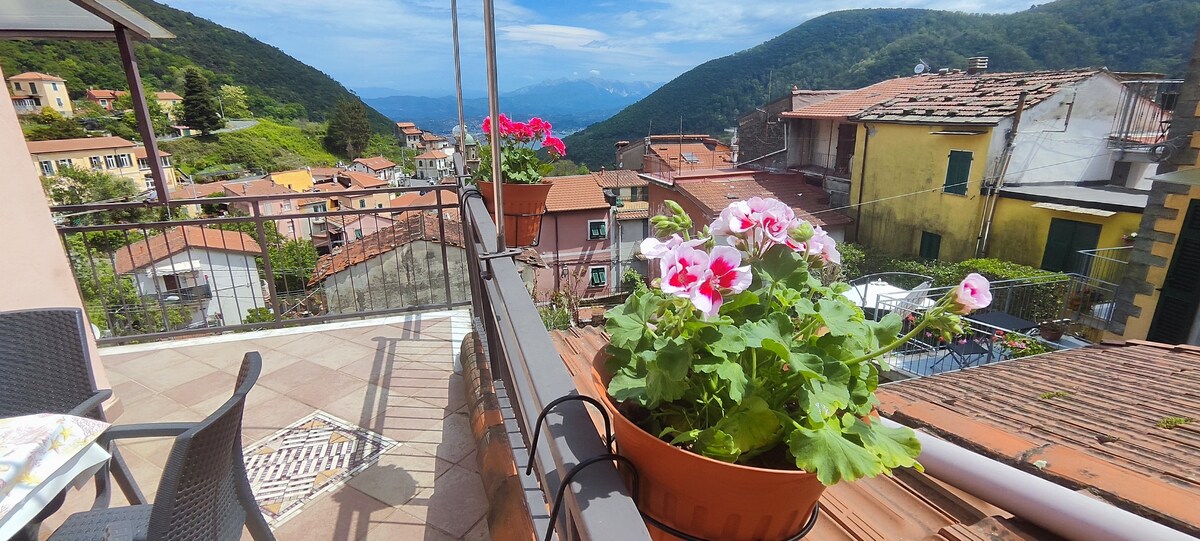A panoramic terrace is adorned with vibrant flowers in pots, overlooking the scenic hills and valleys. The view features rooftops of nearby houses and greenery, with distant mountains visible under a clear sky. A cozy seating area is present for enjoying the outdoors.