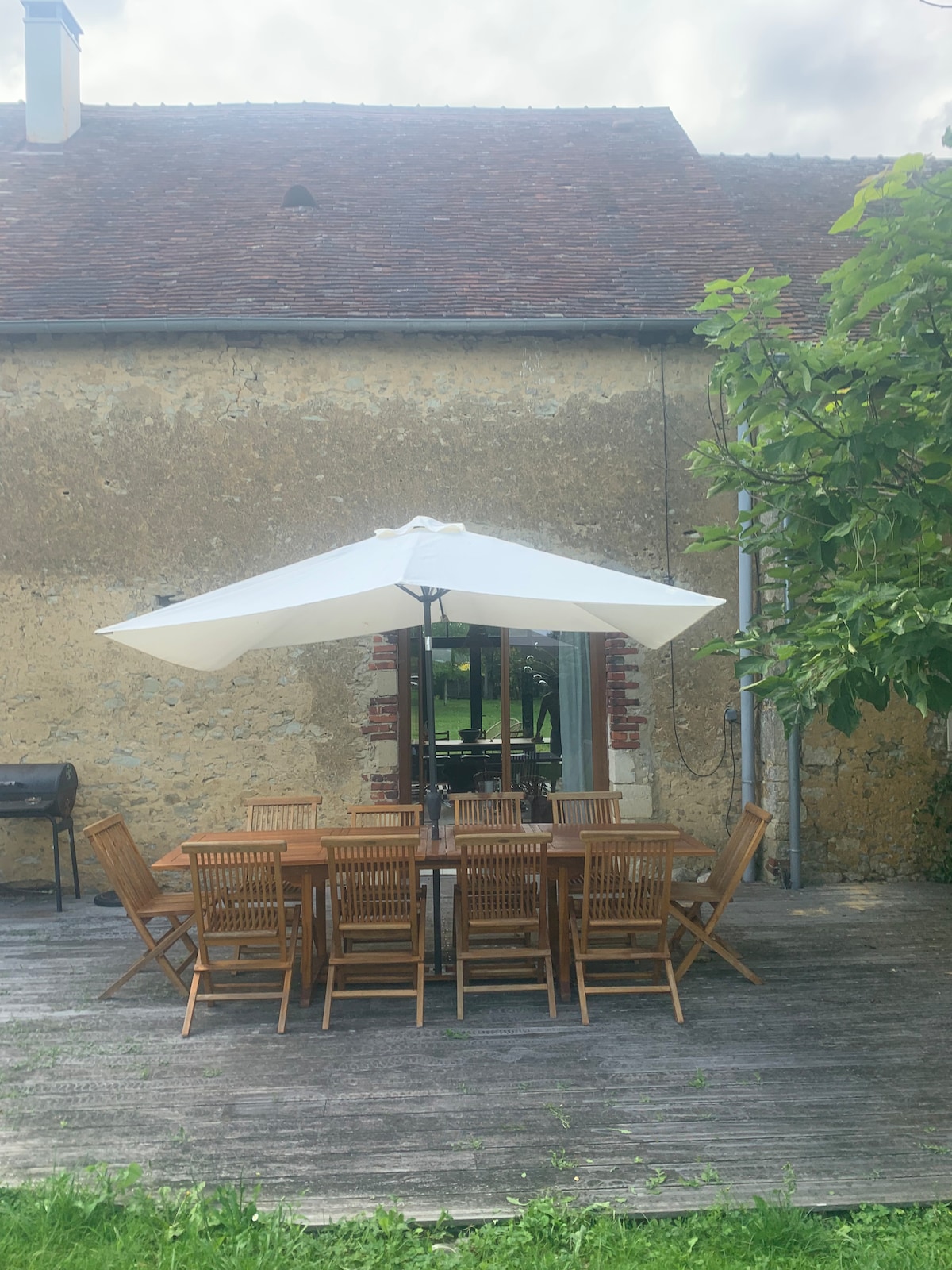 An outdoor dining area is featured with a large wooden table surrounded by multiple chairs, positioned beneath a large white umbrella. A grill is located to the side, and the backdrop showcases a rustic stone wall and greenery.