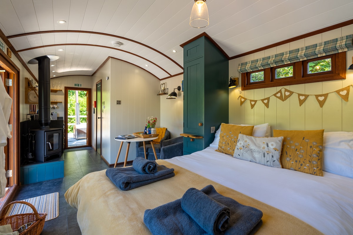 The cozy sleeping area features a spacious king size bed dressed in soft linens, flanked by bedside tables. A wood burner adds warmth, while natural light enters through the windows. A small dining table and chairs create a welcoming nook near the entrance.