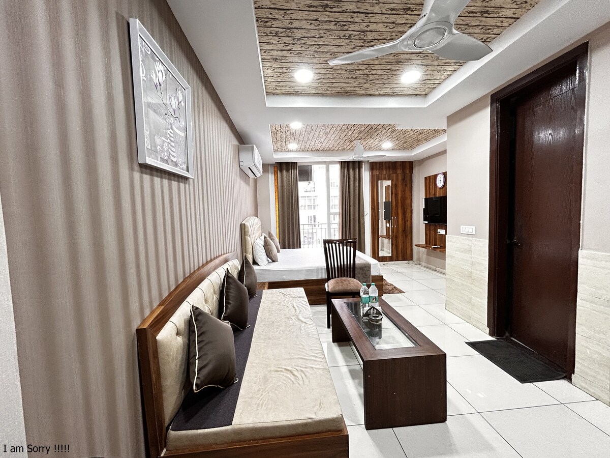 An open living space is presented, featuring a king-sized bed and a wooden sofa with soft cushions. A dining area is visible with a wooden chair and a table. A TV cabinet is situated against the wall, while large windows provide natural light to the room.