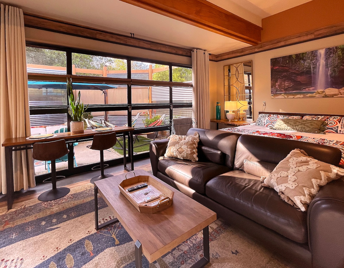 A modern living space features a cozy leather sofa facing a wooden coffee table, complemented by plush cushions. A dining area with two bar stools sits adjacent to panoramic windows, which offer a view of the private courtyard and outdoor lounge area. Natural light fills the room.