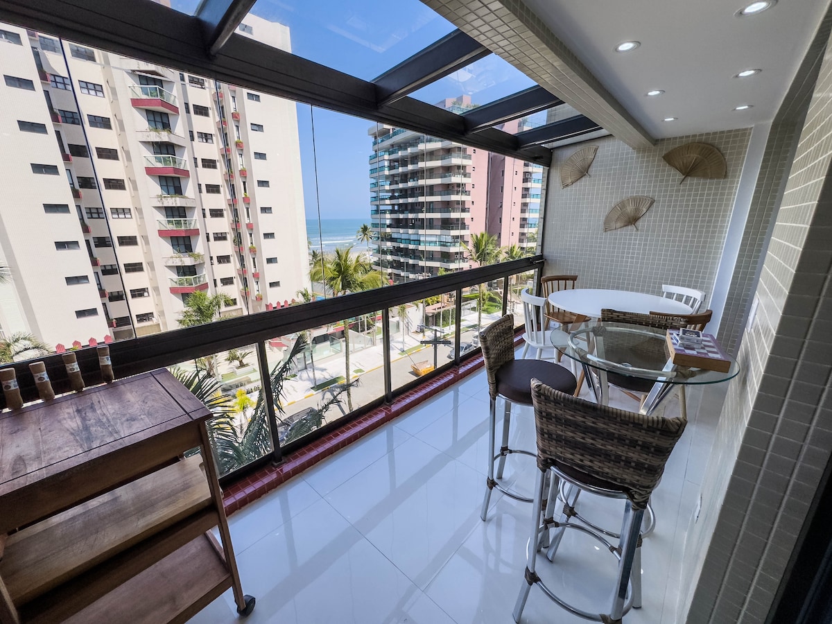 A light-filled balcony with a glass enclosure provides views of the ocean and surrounding buildings. A round glass table is surrounded by wicker dining chairs, and a wooden cart is positioned nearby. The area is enhanced by soft natural light and a tranquil atmosphere.