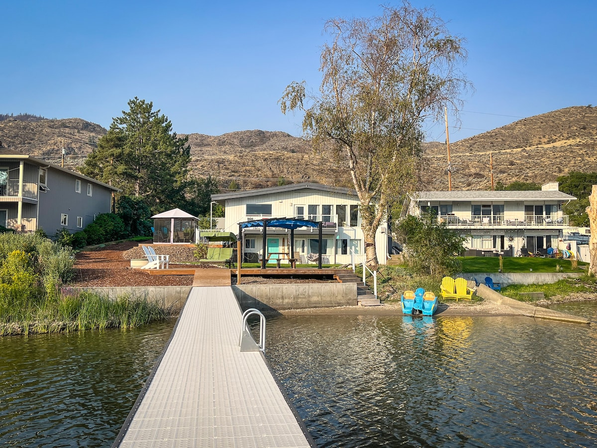 A private dock extends into the water, leading to a spacious lakefront cottage with large windows. Surrounding greenery and landscaped areas complement the setting, while colorful chairs are settled on the shore. Gentle hills can be seen in the background under a clear blue sky.
