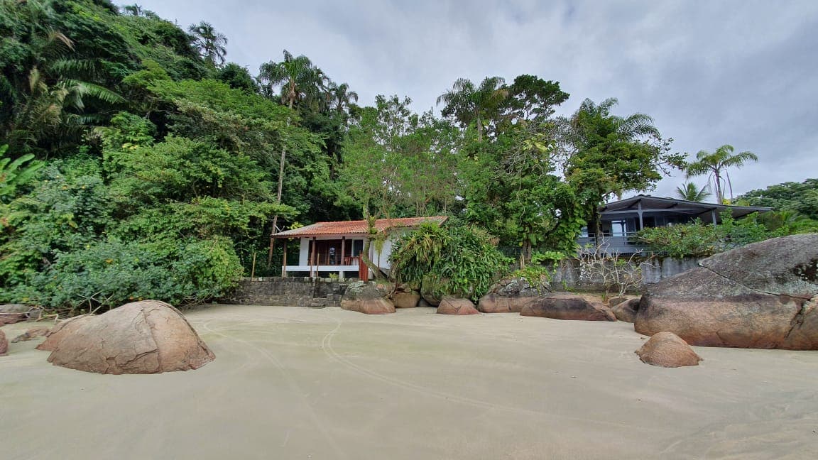 A small house is nestled between lush greenery and rocky formations, situated directly on the sandy beach. The structure features a welcoming porch, surrounded by tropical plants. The coastline offers a serene view of the shoreline, with gentle waves lapping at the sand.