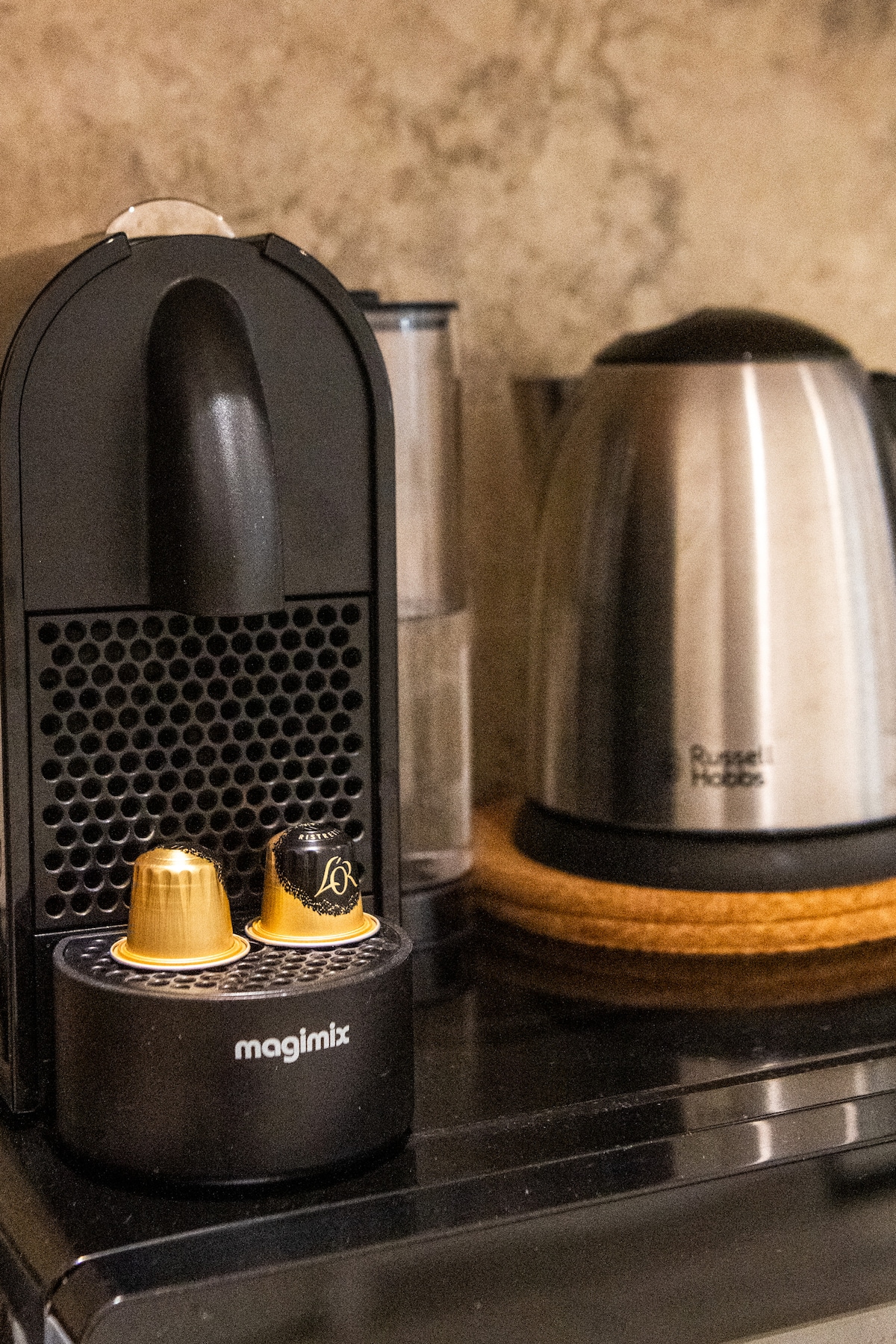 A corner of the kitchen features a Nespresso machine with two capsules placed on its base. Next to it, a stainless steel kettle rests on a wooden trivet, contributing to the functional design of the space.