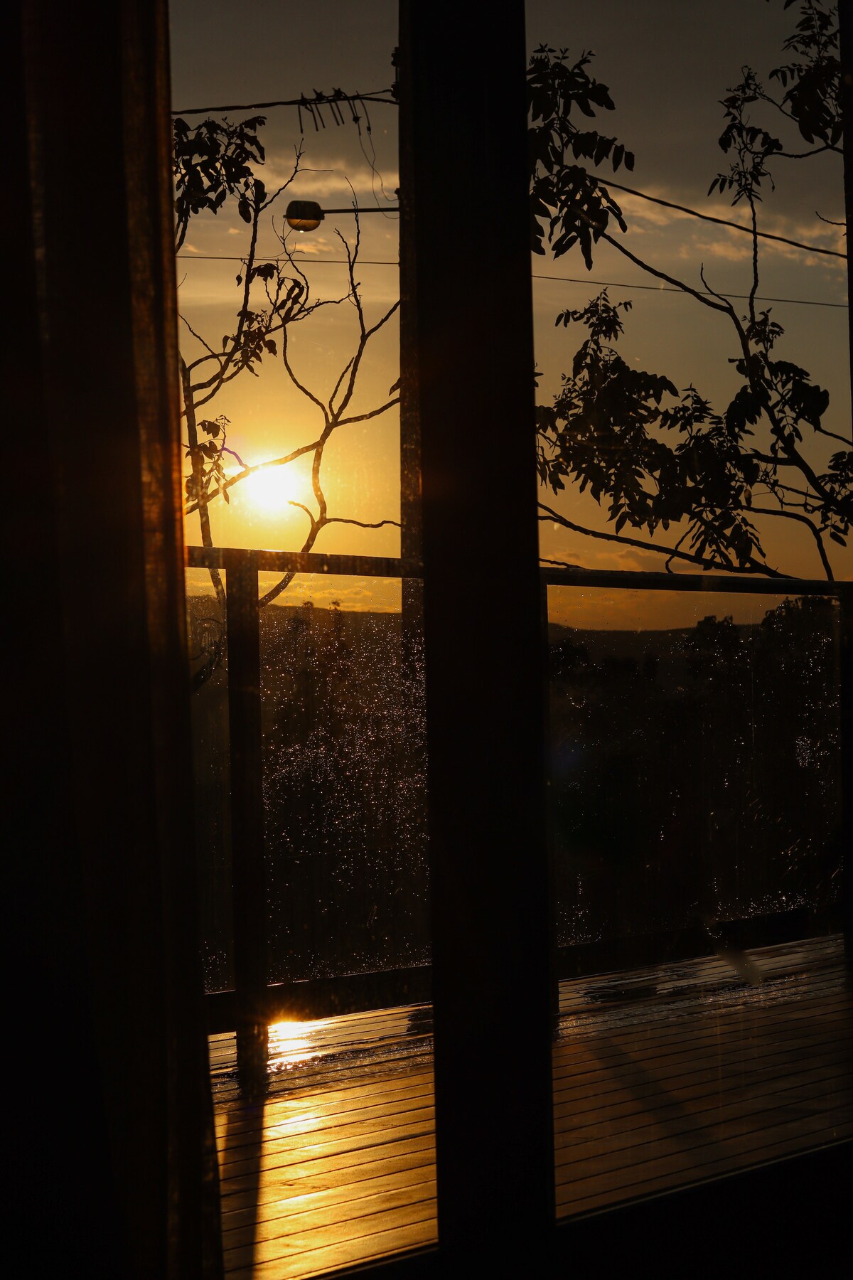 A sunset is captured behind glass doors, creating a silhouette of nearby foliage. The warm glow of the sun reflects off a wooden deck, with raindrops visible on the glass, adding texture to the scene.