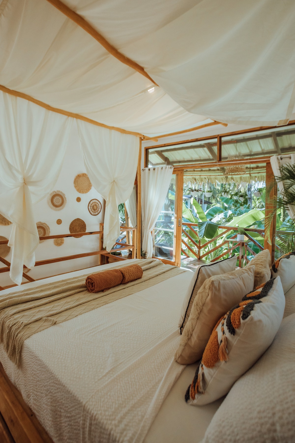 A bright bedroom space is filled with natural light, featuring a spacious bed adorned with neutral bedding. Decorative wall art complements the wooden accents. Large glass doors open to a private balcony, providing views of the lush surrounding greenery.