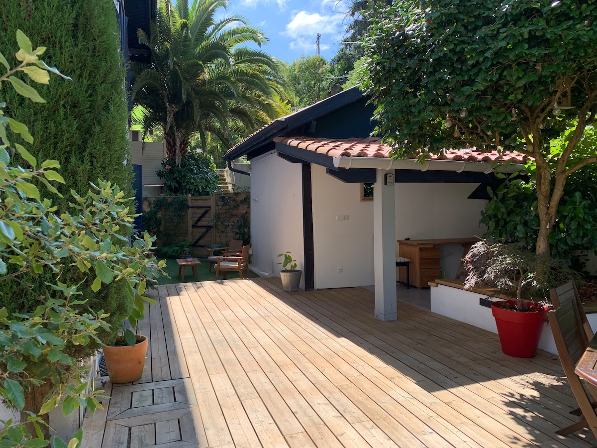 A spacious wooden terrace features a mix of potted plants and seating areas. A covered outdoor space with a tiled roof is seen in the background, surrounded by greenery, adding to the tranquil setting. Natural light illuminates the area, enhancing the inviting atmosphere.