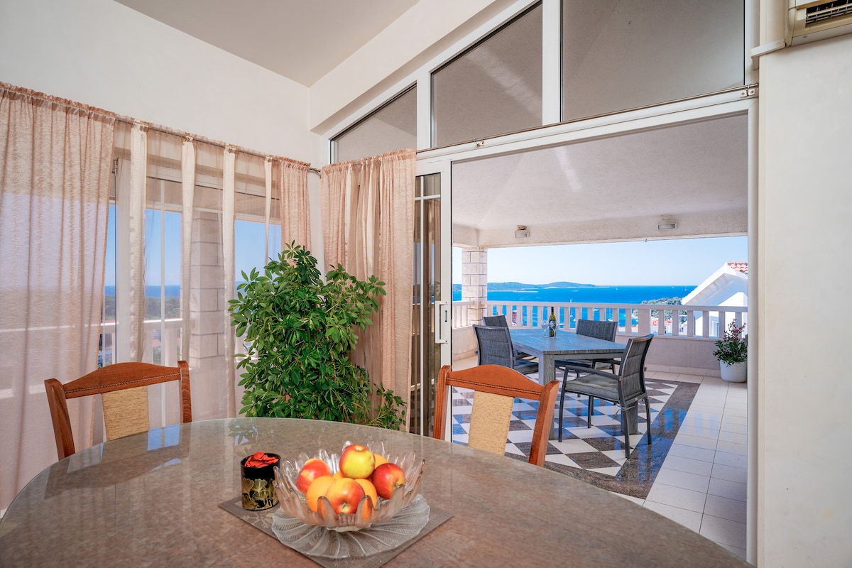 A dining area is presented with a round table featuring a fruit bowl at its center. Large glass doors open to a terrace, allowing natural light to fill the space while offering views of the sea and surrounding landscape. Soft curtains gently frame the windows.