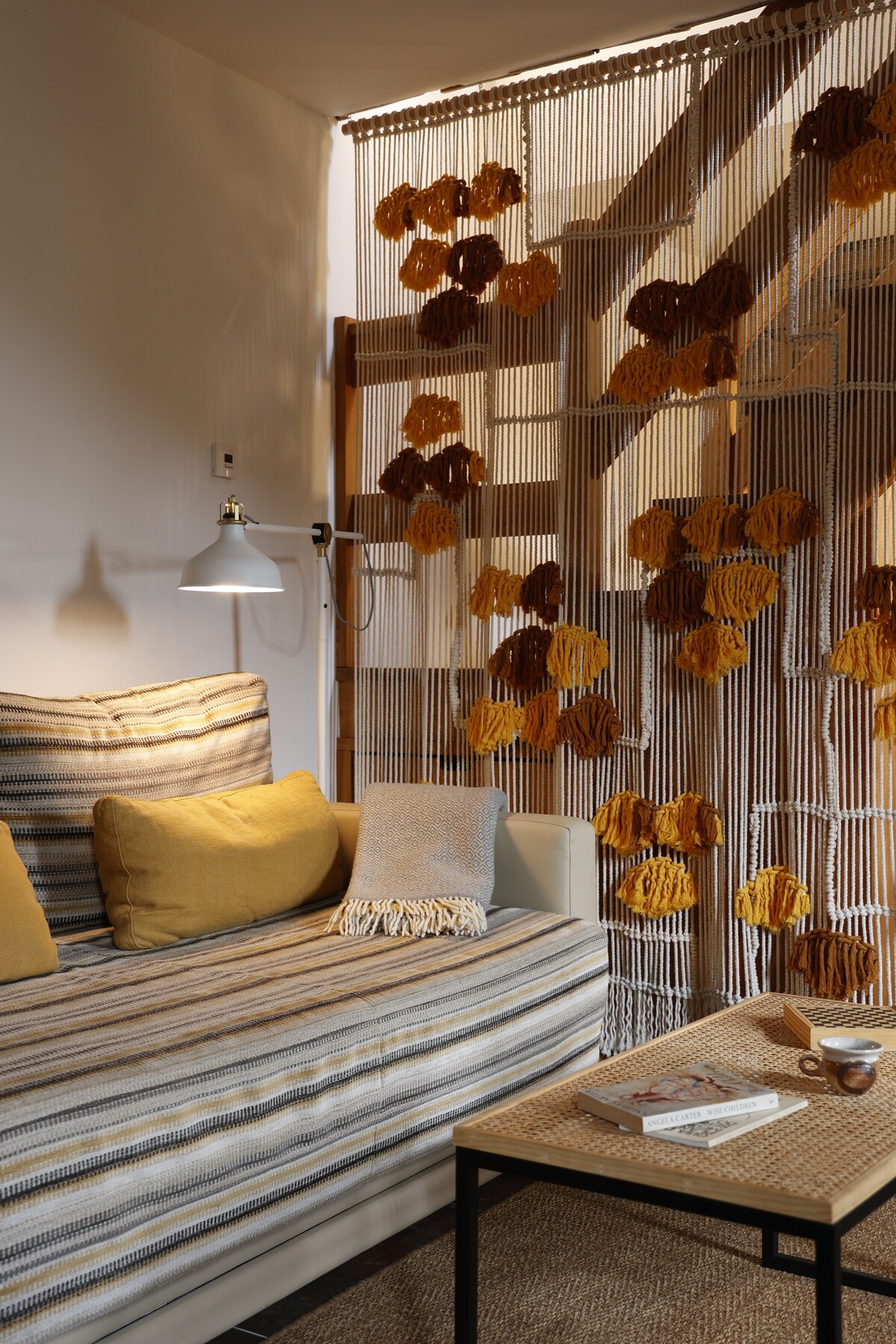 A cozy seating area features a striped sofa adorned with yellow throw pillows. A woven macramé screen hangs nearby, decorated with flowers in warm tones, complementary to the inviting natural light that fills the space.