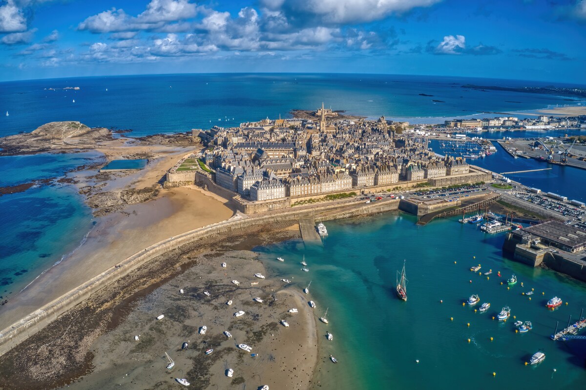 An aerial view presents the historic city of St Malo bordered by the sea. The coastline features sandy beaches and a marina filled with boats. Stone structures and fortifications are visible, creating a contrast against the blue waters and sky.