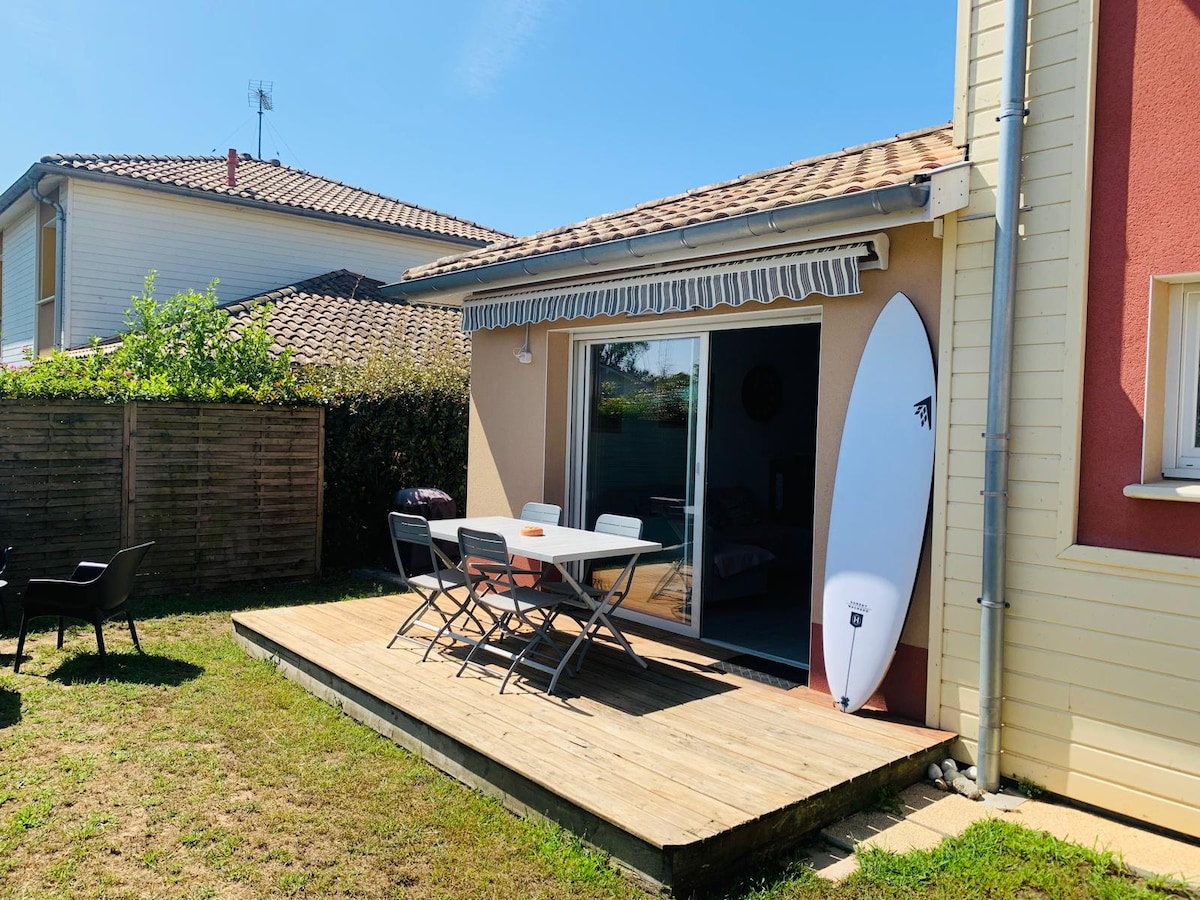 A wooden deck extends from the house, featuring a dining table with chairs. A surfboard leans against the wall, and sunlit grass surrounds the area. Sliding glass doors provide access to the interior, while potted plants and a privacy fence enhance the outdoor setting.