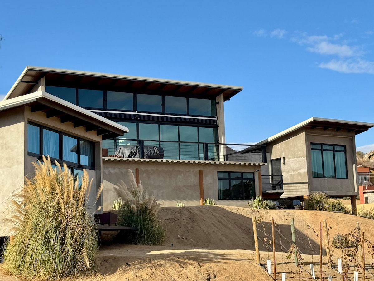 A modern architectural structure features large windows that provide natural light and views of the surrounding landscape. The two interconnected buildings are complemented by desert vegetation and a sandy area, emphasizing a harmonious relationship with the environment.