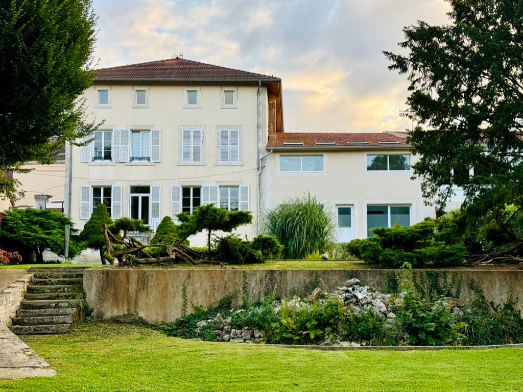 The exterior of the spacious house features large windows and multiple balconies, surrounded by manicured gardens. A grassy area is visible in front, with stone steps leading up to the house. The serene sky is tinged with soft hues of sunset.