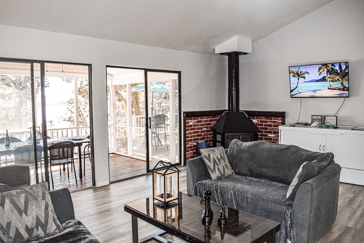 The living area features comfortable gray sofas, a sleek coffee table, and a flat-screen TV mounted on the wall. Large sliding glass doors provide access to the outdoor porch, while a modern fireplace adds to the cozy ambiance.