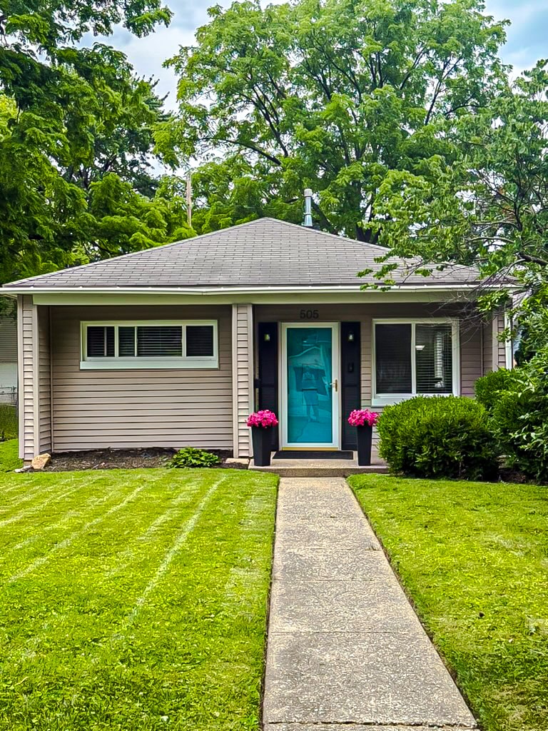 A charming exterior view of the single-story home is presented, featuring a well-maintained lawn and a concrete pathway leading to a bright blue front door. Flanking the entrance, vibrant pink flowers add a touch of color, while lush green trees provide a natural backdrop.