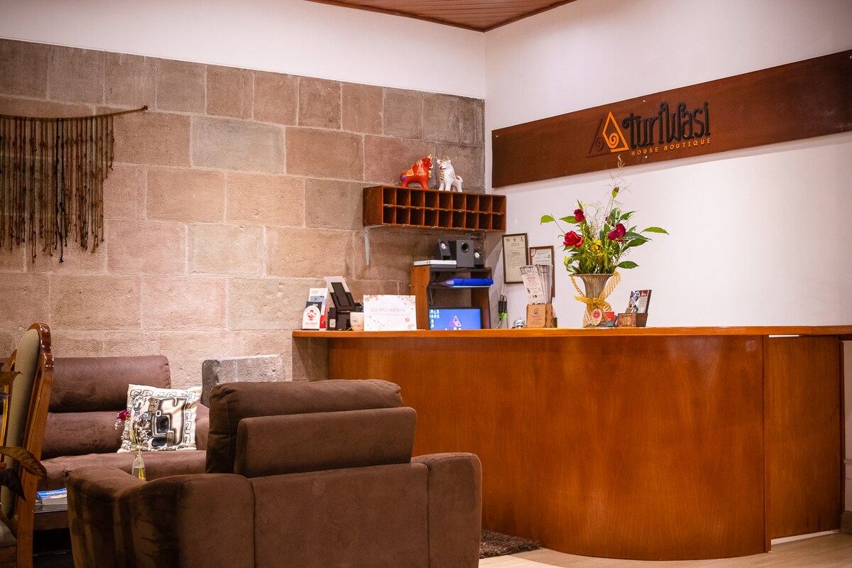 A reception area is featured with a warm wooden desk and a friendly layout. Decorative plants and local artwork are arranged on the desk, while comfortable seating is provided nearby. Stone walls add texture to the space, creating a welcoming environment for guests.