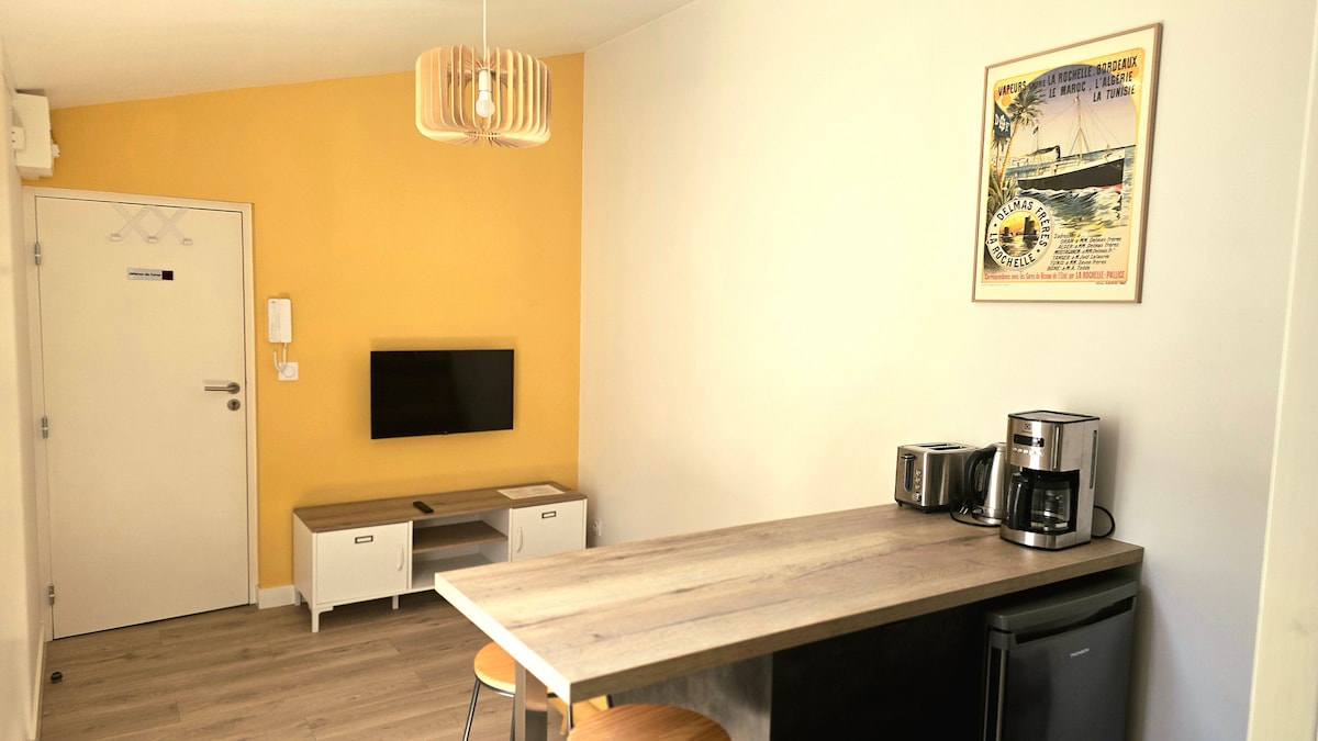 A modern living area features a bar counter with two stools, complemented by a compact refrigerator and coffee-making appliances. A flat-screen TV is mounted on the wall, while a framed art piece adds a pop of color. The door leads to the corridor.