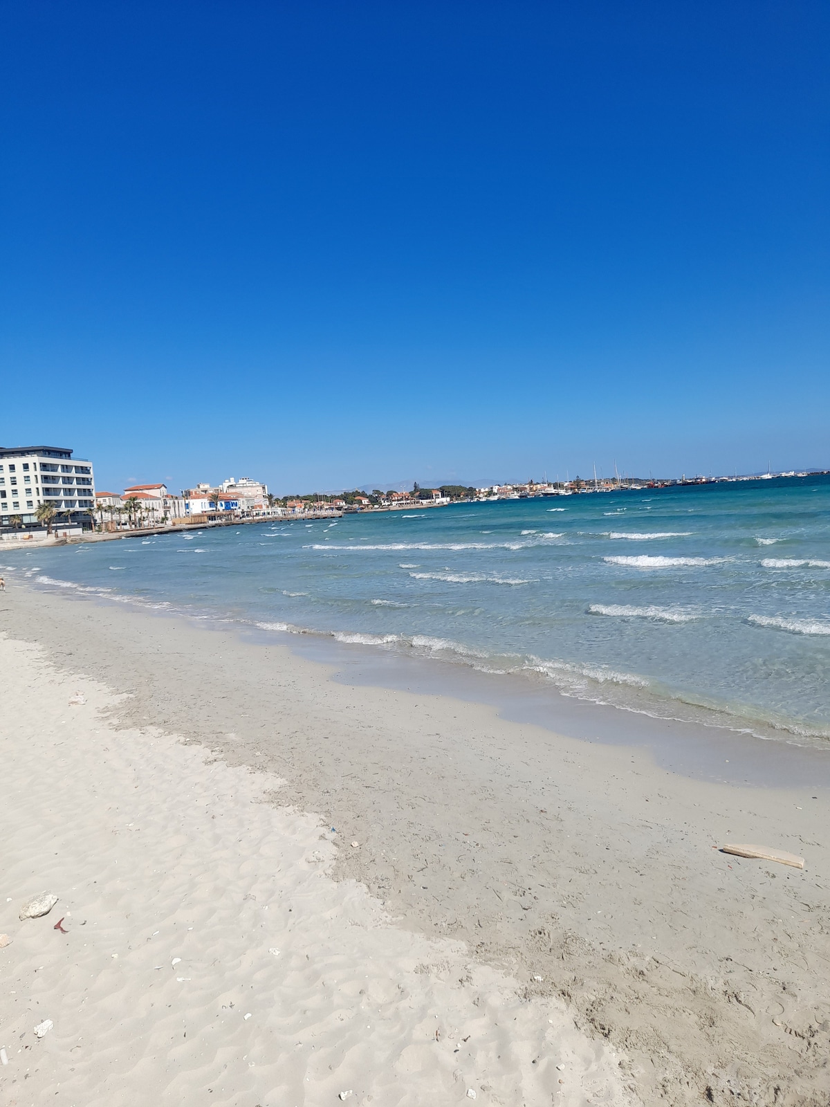 A sandy beach stretches along the shoreline, gently meeting the turquoise waves. In the distance, a waterfront city is visible, with buildings contributing to the coastal view. Clear blue skies overhead enhance the serene beach atmosphere.