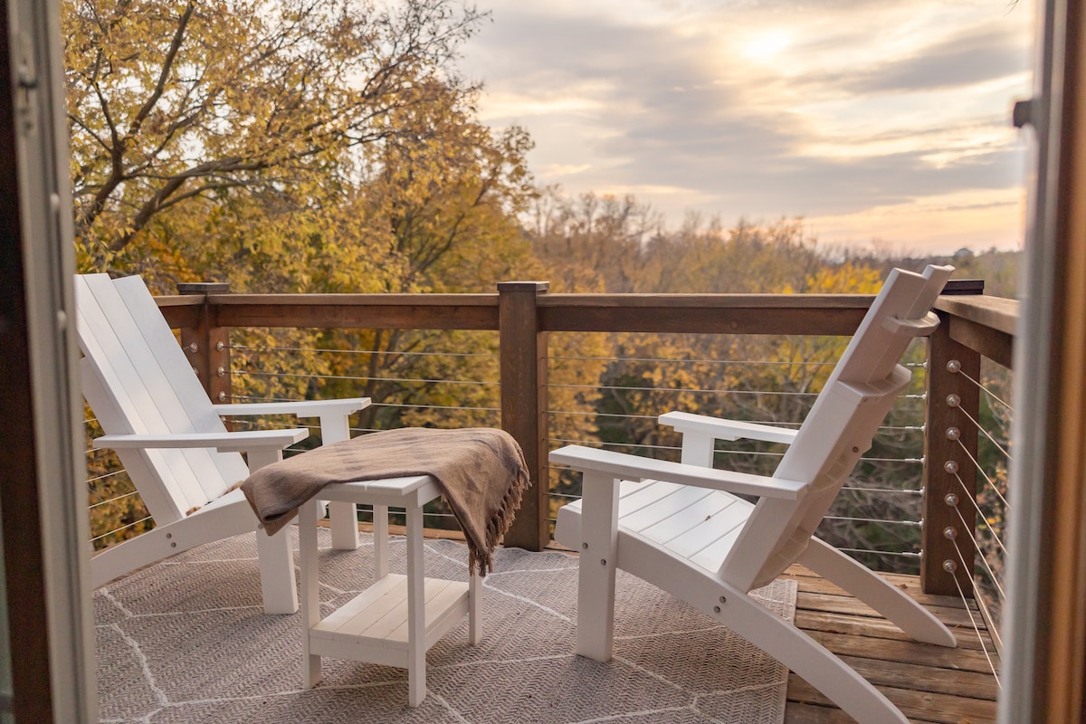A serene outdoor seating area features two white Adirondack chairs positioned on a wooden deck. A small table sits between them, with a soft brown blanket draped over one chair. The backdrop showcases vibrant autumn trees under a softly lit sky.