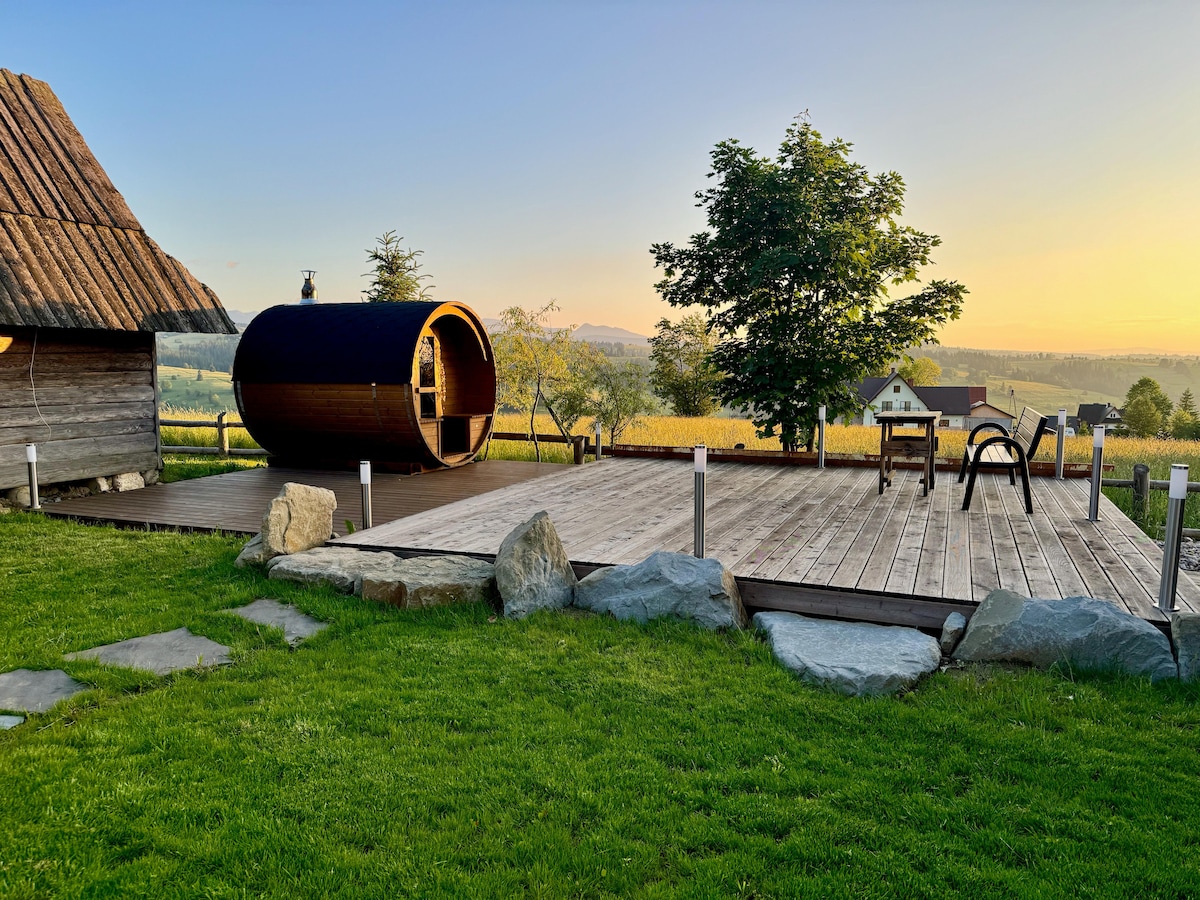 An outdoor area is featured, showcasing a wooden deck surrounded by natural stone. A cylindrical sauna is prominently positioned beside the deck, with a view of lush greenery and rolling hills in the background. The scene is illuminated by warm evening light.