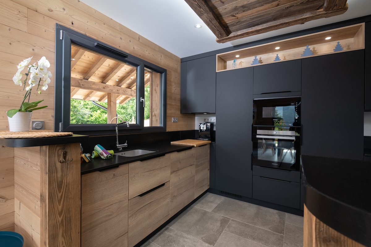 The modern kitchen features sleek black cabinetry paired with wooden accents, offering a harmonious blend of materials. A stainless steel oven and cooktop are included, complemented by a stone countertop. Natural light enters through large windows, providing views of the surrounding nature.