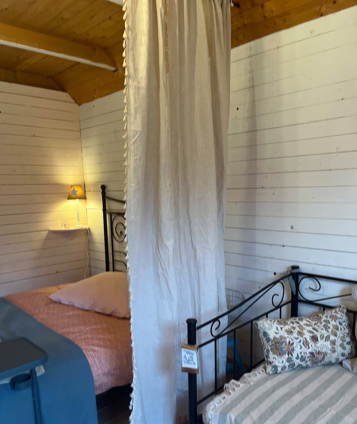 The room features two beds separated by a soft fabric divider. One bed is adorned with a light pink pillow and a pastel coverlet, while the other has a striped blanket and decorative pillows. Soft lighting from a nearby lamp enhances the tranquil setting.