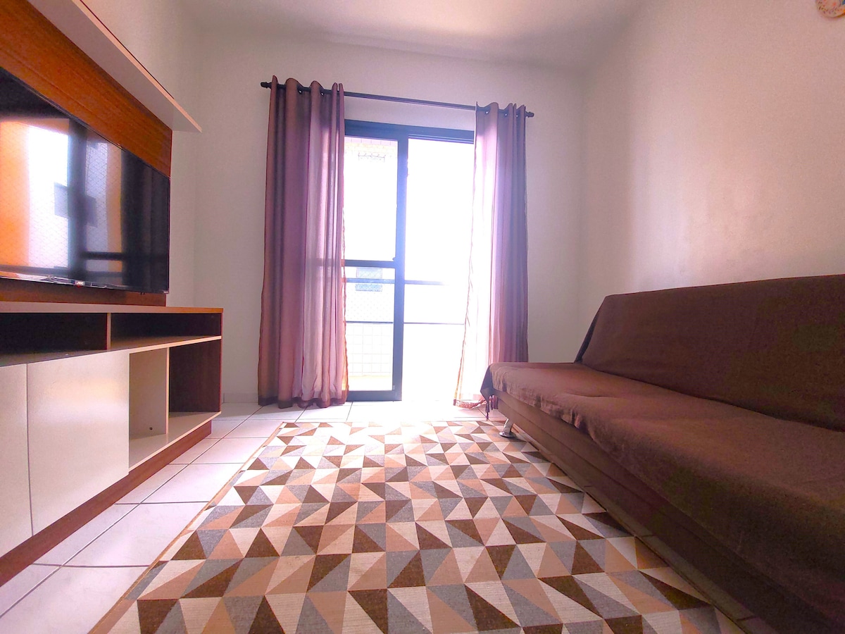 A comfortable living space features a brown sofa bed positioned against a modern entertainment unit that holds a flat-screen television. Large windows are dressed with sheer curtains, allowing natural light to brighten the room. A geometric patterned rug covers the tiled floor.