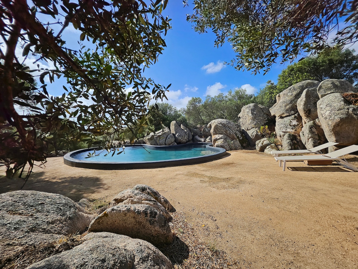 A secluded pool area is nestled among natural boulders and Mediterranean vegetation. The circular pool features a smooth edge, surrounded by a sunbathing area with white loungers, providing a serene spot for relaxation. The sky above is clear with soft clouds.