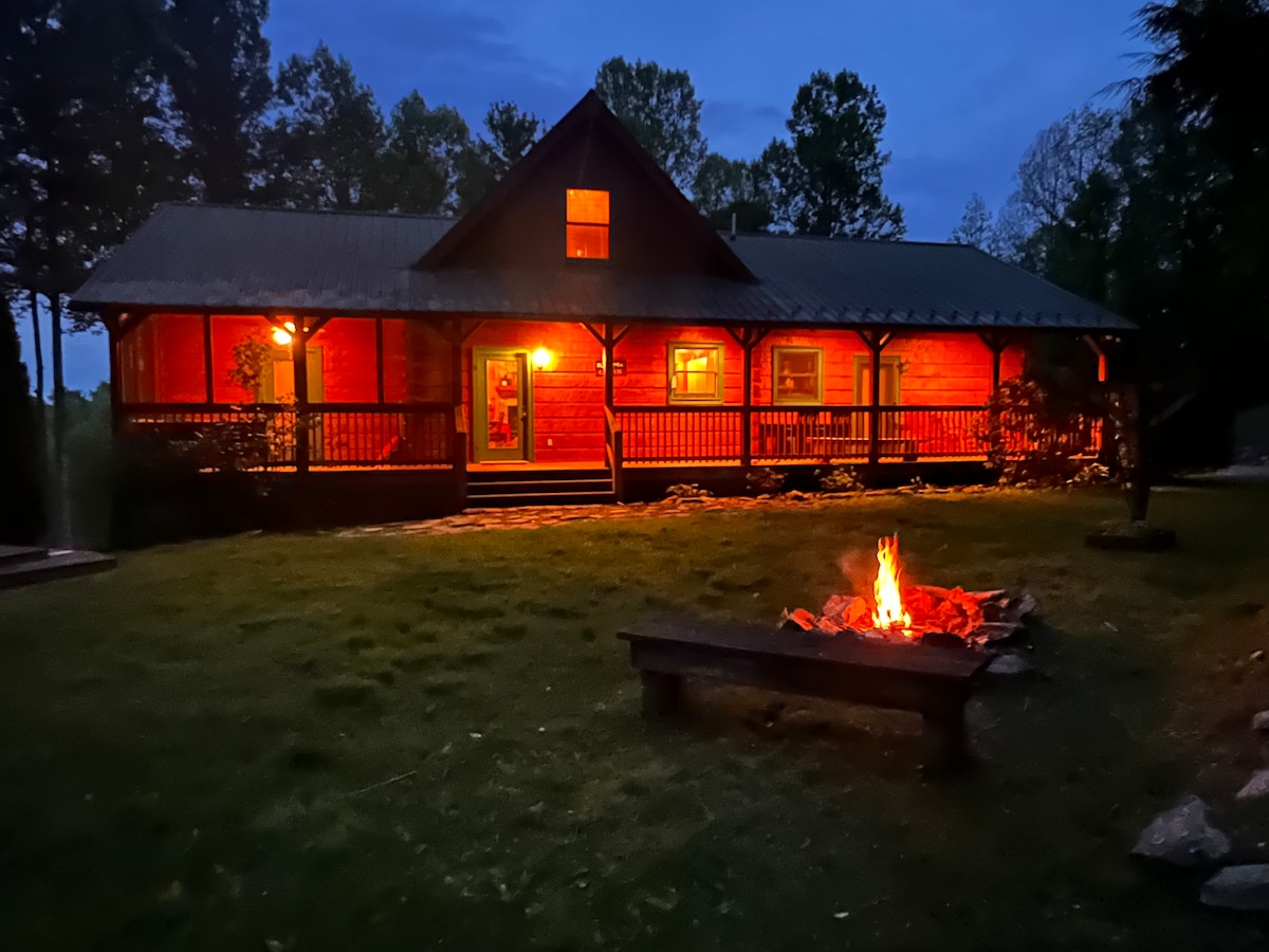 A cozy cabin is illuminated in warm light, set against a twilight sky. A fire pit with a crackling fire is placed on a grassy area in front of the cabin, surrounded by stones. The inviting porch features ample seating and rustic wooden beams.