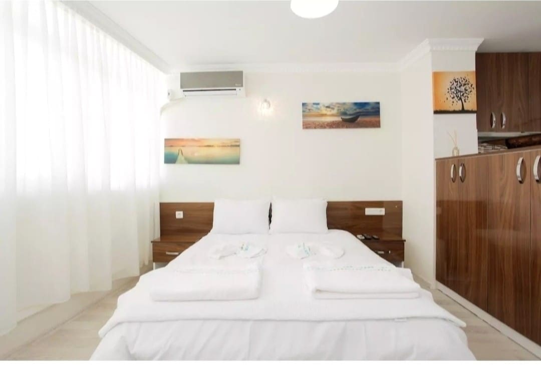 A spacious sleeping area is featured, with two neatly arranged beds topped with fresh white linens and towels. Soft natural light filters through sheer white curtains. A simple wooden cabinet is visible in the corner, along with colorful artwork adorning the walls, enhancing the serene ambiance.