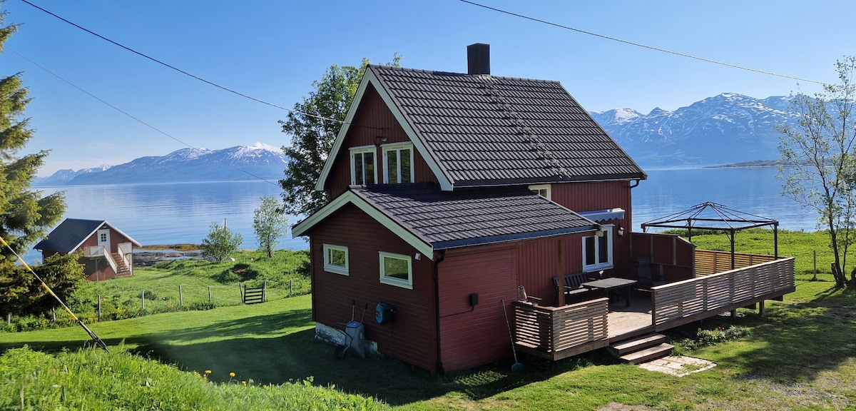 The red wooden cabin is set against a backdrop of clear blue waters and snow-capped mountains. A spacious deck extends from the cabin, featuring outdoor seating. Lush greenery surrounds the property, and a distant building is partially visible, adding to the serene landscape.