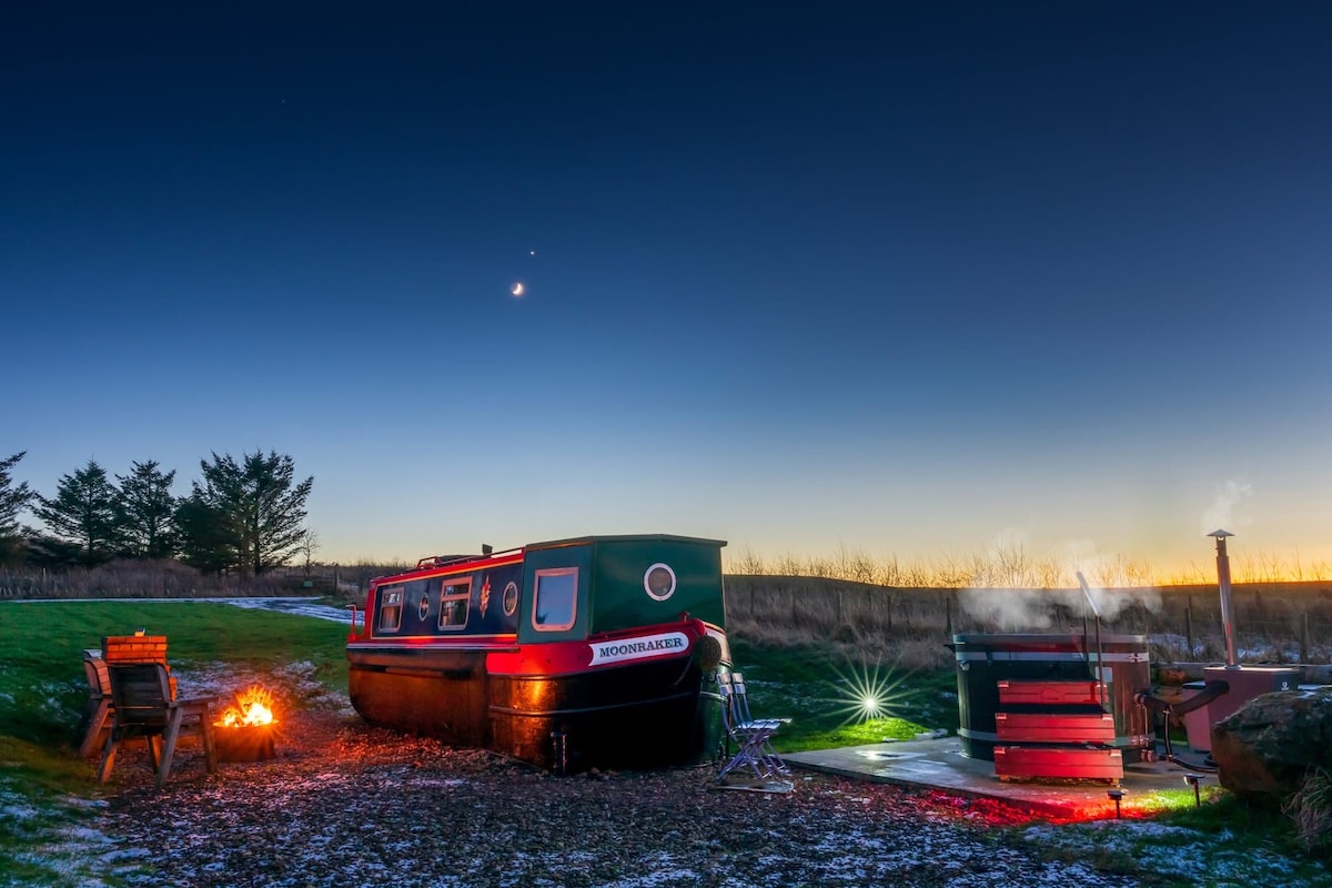A narrowboat named Moonraker is positioned on a grassy area, illuminated by the evening sky. A fire pit and barbecue grill are situated nearby, with garden chairs arranged for relaxing. The background features trees against a gradient sky transitioning from blue to orange.