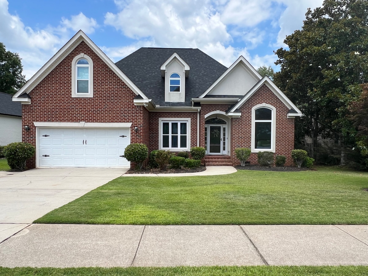 A spacious two-story brick home is presented, featuring a well-manicured lawn and clean concrete driveway. The front entrance includes a covered porch with two steps leading to the main door. Large windows allow natural light to fill the interior, framed by neatly trimmed shrubs.