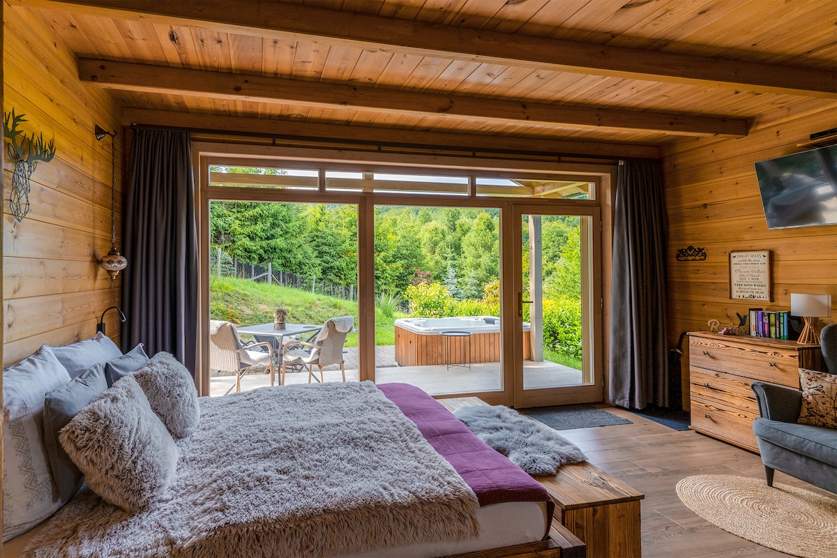 A spacious bedroom features a comfortable bed with plush bedding and generous pillows. Large glass doors provide access to an outdoor area with a jacuzzi and seating. Natural light spills in, illuminating the wooden interior and creating a serene retreat.