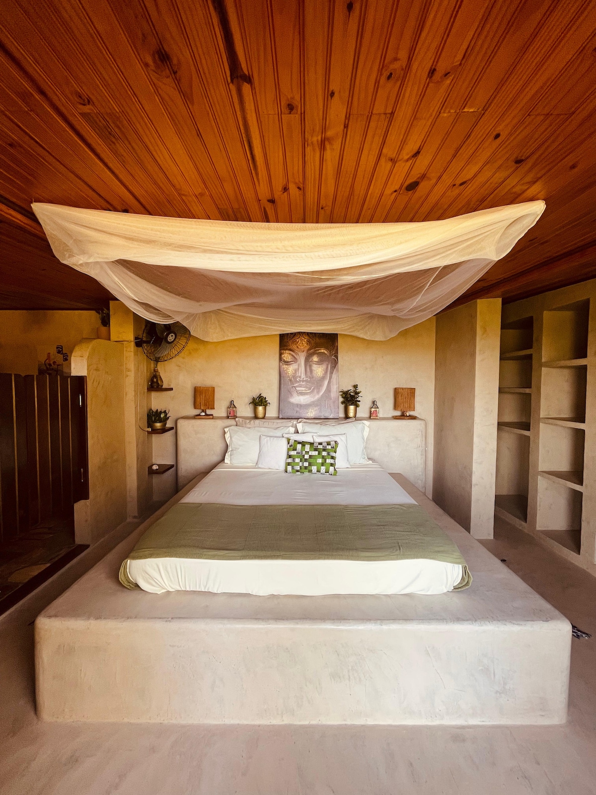 The sleeping area features an expansive bed adorned with a light green blanket and complemented by a decorative pillow. Natural wood ceilings enhance the serene atmosphere, while built-in shelving provides storage. Subtle indoor plants and soft lighting elements introduce a calming touch to the space.