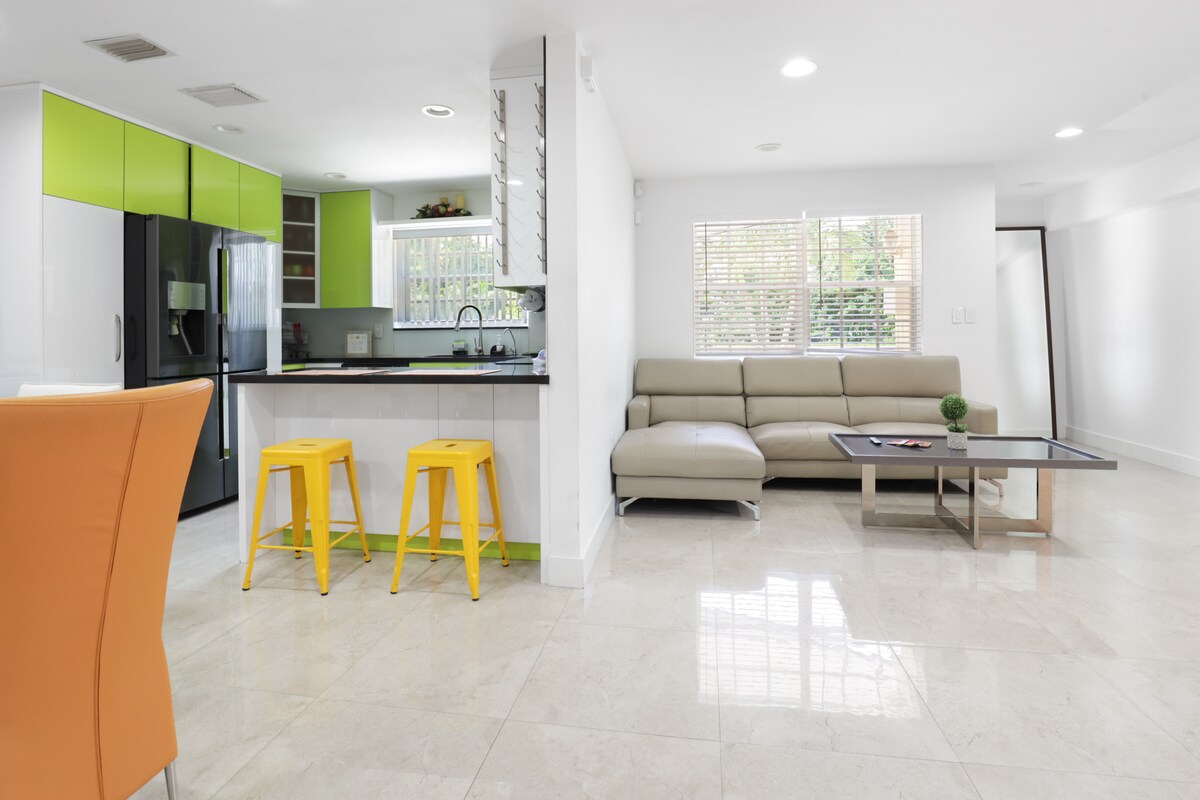 A modern living area features a sleek beige sectional couch paired with a stylish coffee table. Bright yellow bar stools accent a contemporary kitchen with vibrant green cabinetry. Natural light streams through the window, highlighting the polished stone flooring.
