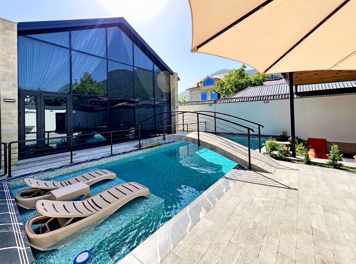 A tranquil private pool area features a wooden bridge leading over clear water. Lounge chairs are positioned at the pool's edge. Sunlight reflects off the water, while a covered seating area provides shade. Lush greenery and a garden enhance the serene outdoor environment.