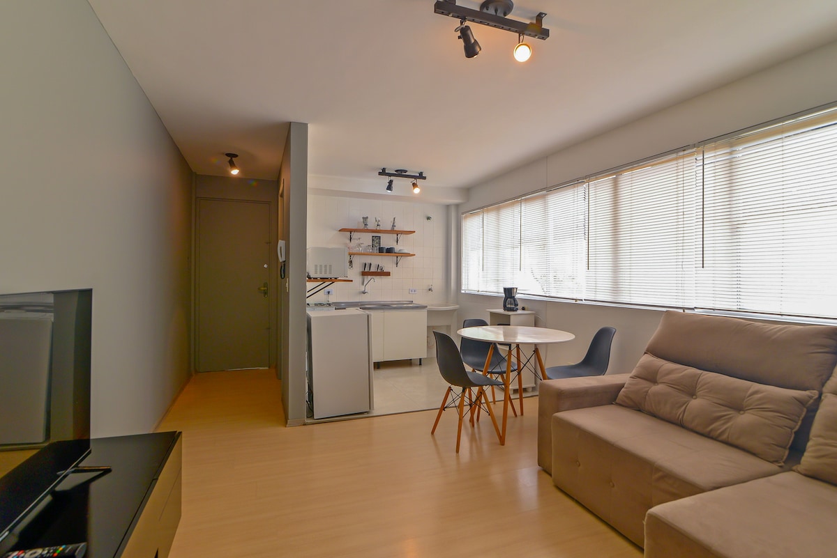 An open living space features a comfortable sofa bed and a round dining table with four chairs. Large windows provide natural light, while the kitchen area showcases modern appliances. Light fixtures on the ceiling create a warm ambiance, contributing to the overall functional layout.