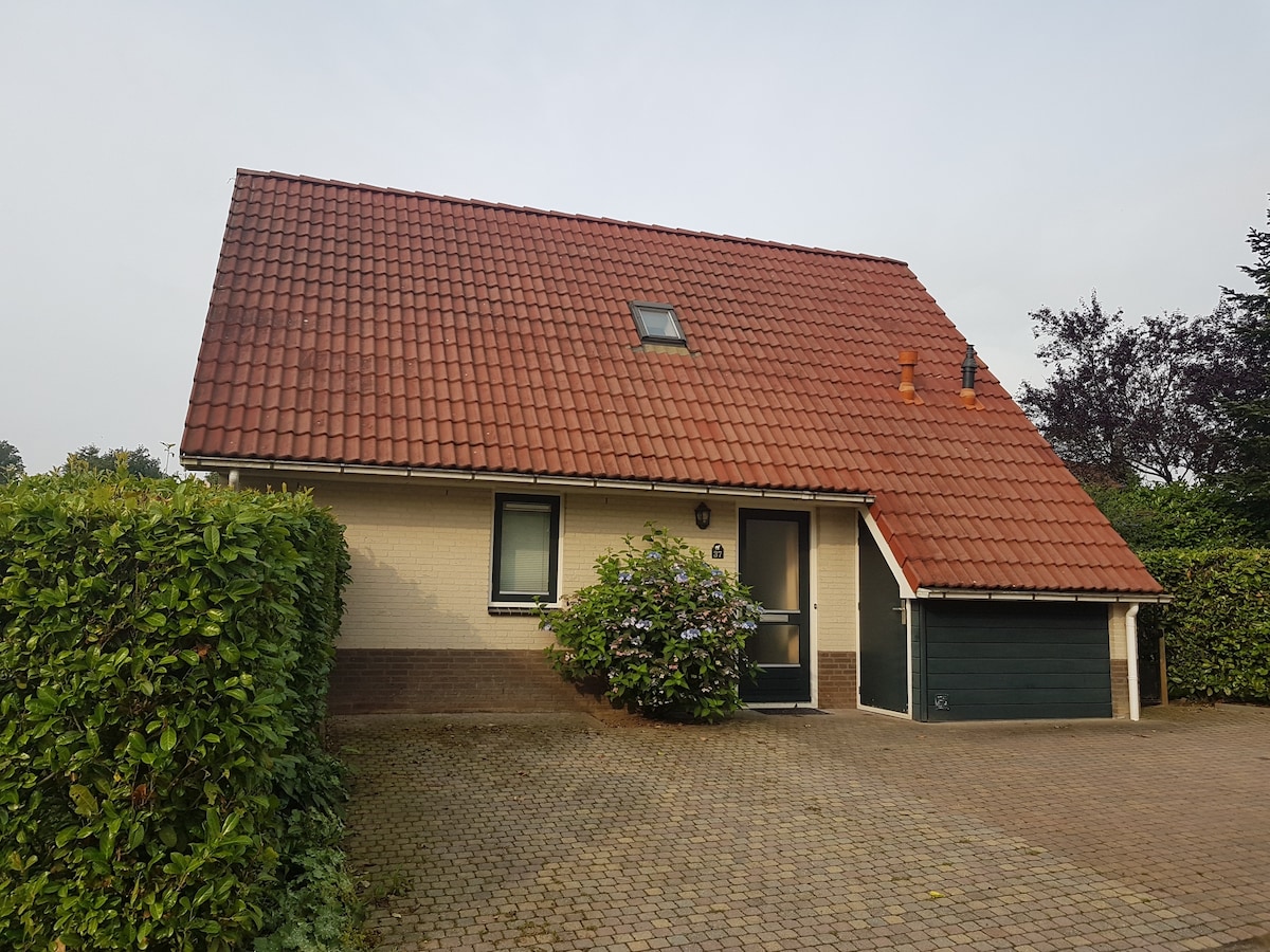 A freestanding vacation home is set within a garden, featuring a red-tiled roof and a front door framed by greenery. A driveway made of cobblestones leads up to the entrance, with a garage visible on the right side of the house.